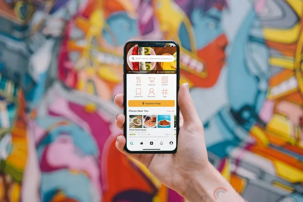 Mão segurando um smartphone que exibe a interface do aplicativo "ContaPraGente", mostrando opções como busca por pratos, restaurantes e produtos, com um vibrante mural colorido ao fundo.