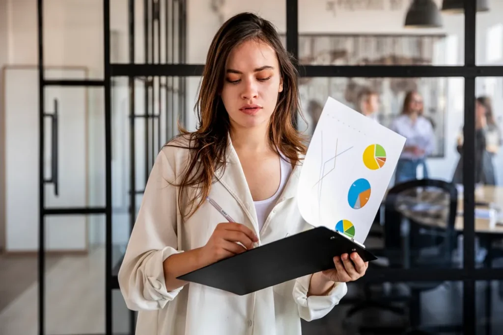 Profissional segurando prancheta com gráficos de pizza e linha, conferindo resultados de negócios em ambiente de escritório com vidro e colegas ao fundo