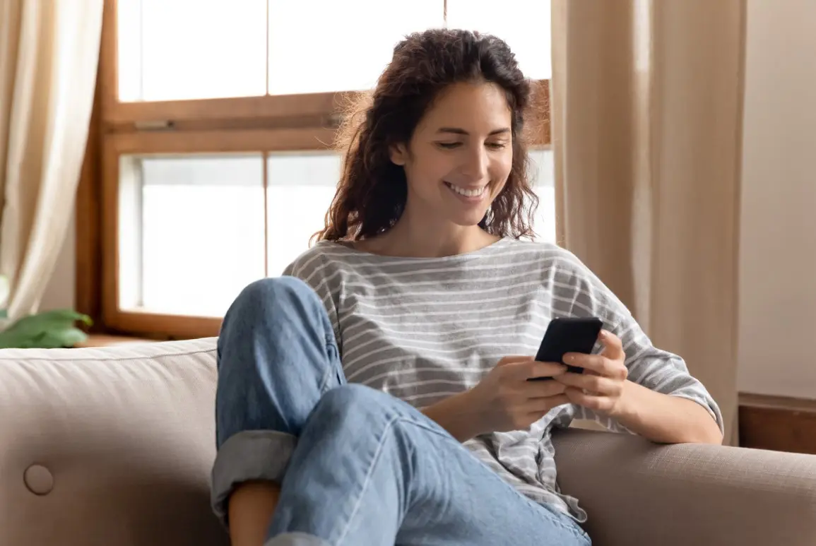 Uma mulher branca, de cabelo castanho ondulado, está usando uma blusa cinza com listras brancas e calça jeans azul claro. Ela está mexendo no telefone sorrindo porque está recebendo atendimento de sua loja pelo telefone. Isso é possível graças à estratégia de ominichannel que a loja implementou