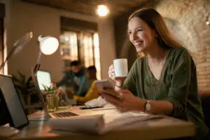 Feliz empresário usando telefone e bebendo café enquanto trabalhava até tarde em seu escritório e acompanhando pelo celular sua entrega..