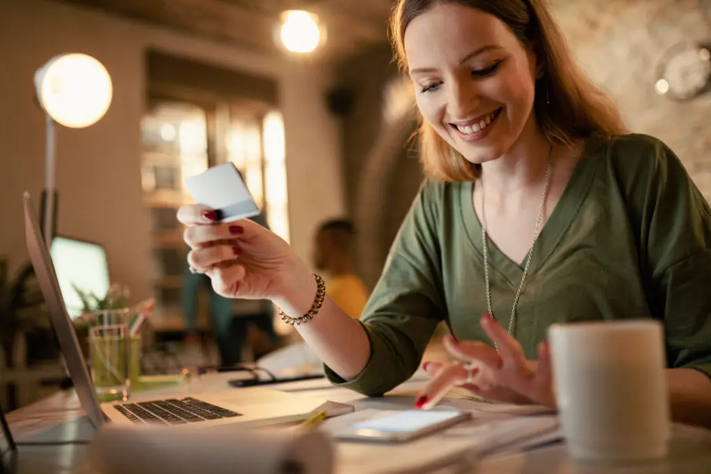 Mulher de negócios feliz usando smart phone e cartão de crédito enquanto fazia compras na Internet à noite no escritório. Ela irá acompanhar sua entrega pelo celular pois a empresa utiliza o Last Mile, onde o cliente acompanha a rota do pedido.