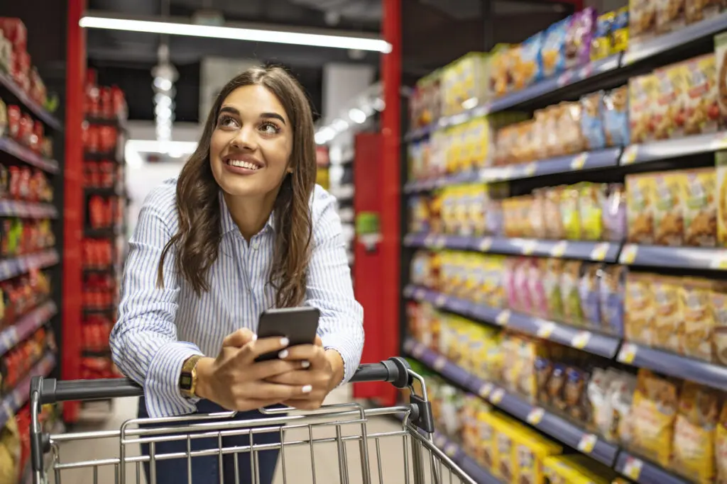 Mulher sorridente no supermercado. Mulher feliz no supermercado. Mulher jovem e bonita às compras em uma mercearia / supermercado. Listas de compras em formato de aplicativo.