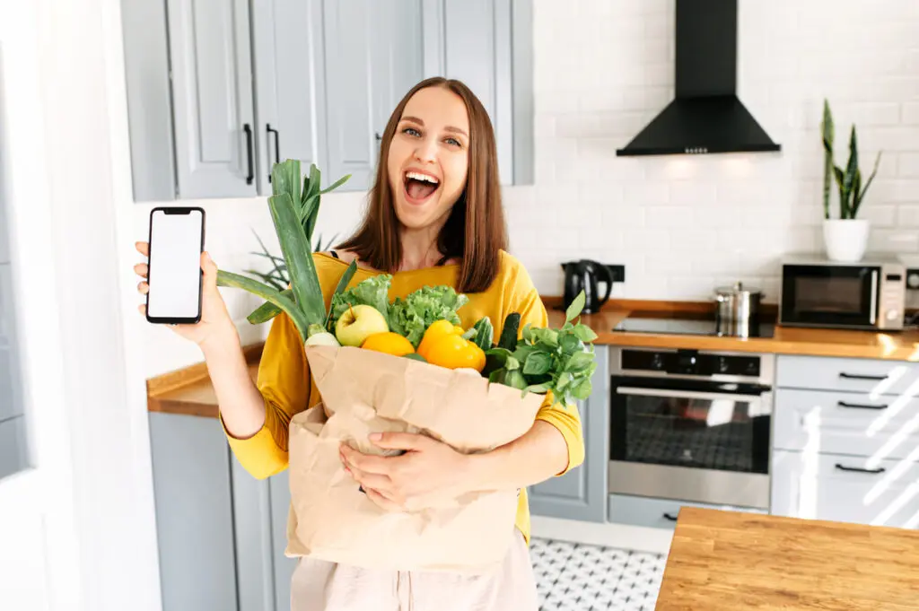 Mulher jovem feliz com um saco de papel cheio de vegetais frescos ecológicos fica na cozinha e mostra o telefone com tela em branco. App para entrega de produtos agrícolas.