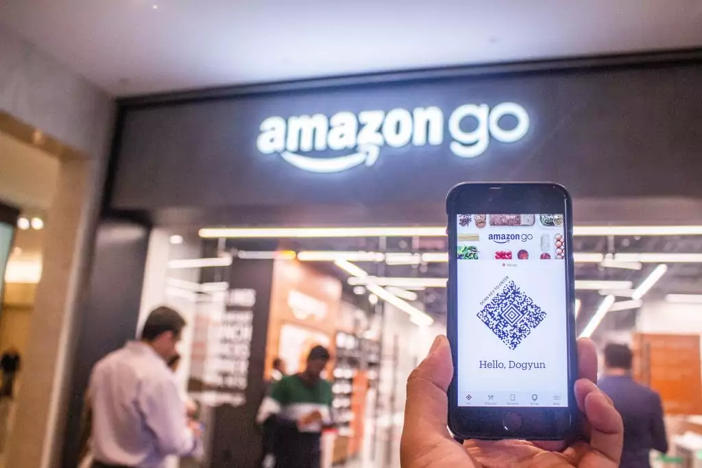 Imagem de mão segurando o celular em frente a Amazon Go. Visual merchandising, e sua relação com a experiência de compra do cliente.
