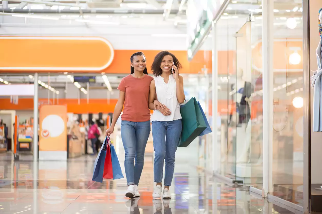 Duas mulheres negras estão carregando sacolas de compras, uma está conversando no celular enquanto a outra olha para a vitrine.