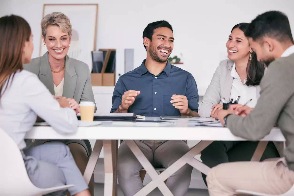 Um grupo de pessoas está sentado em uma mesa conversando e rindo enquanto trabalham. O time está unido e engajado mostrando a eficácia da estratégia de Gestão de Pessoas