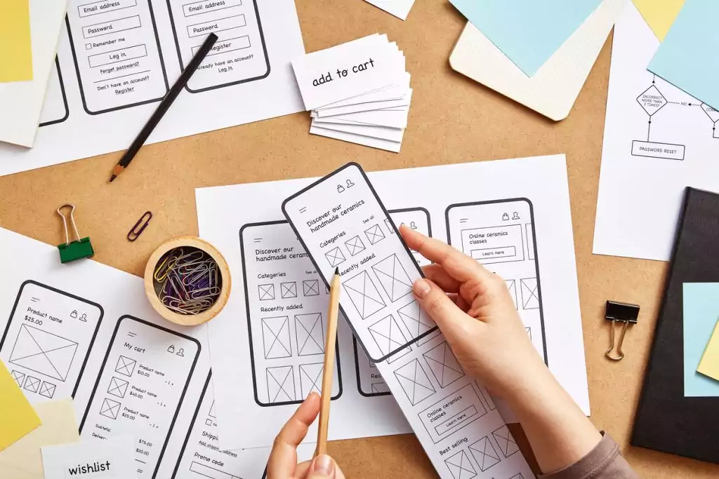 Uma mesa está cheia de papéis com desenhos de páginas de internet e alguns lápis. Uma mulher está segurando uma folha e planejando qual a melhor interface a ser escolhida.