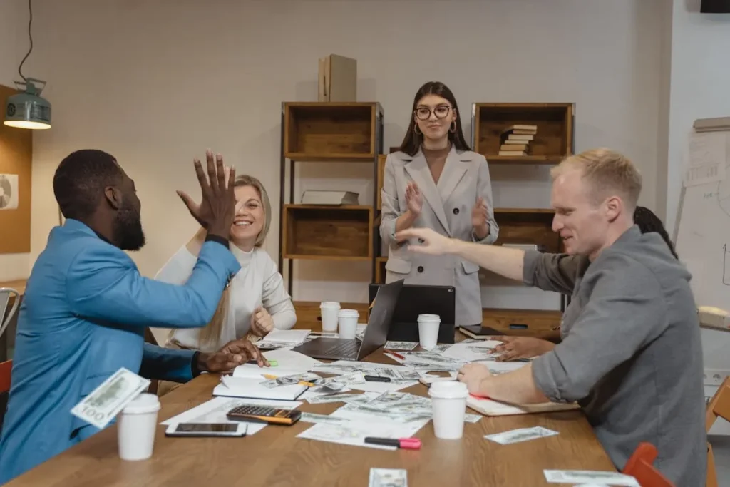 Equipe de gestão celebrando sucesso em reunião de escritório, com dois homens fazendo um "high-five" e uma mulher aplaudindo, simbolizando gestão de pessoas eficaz e recompensa financeira.