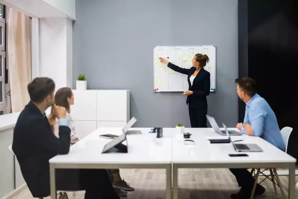 Uma equipe está reunida em uma sala realizando uma reunião. Uma mulher de roupa social preta está apontando para um quadro dividido em etapas de conclusão de um projeto. A mulher está mostrando a gestão de projetos que ela elaborou para o serviço