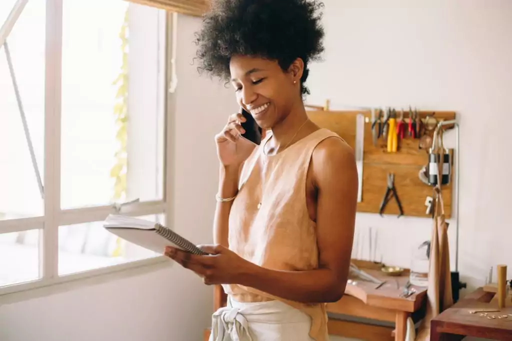Uma mulher negra de blusa bege e calça brance está em pé conversando no telefone. Ela sorri e olha para baixo, na direção de um caderno que segura em suas mãos contendo detalhes do pedido de um cliente. Ela está aumentando as vendas por meio do uso do WhatsApp Business