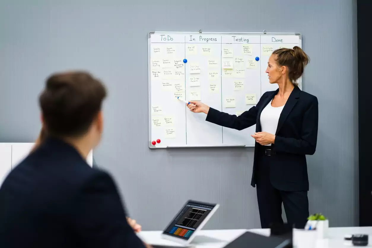 Uma mulher de roupa social preta e coque está em pé apresentando a gestão de projetos que ela elaborou para guiar a sua equipe de trabalho