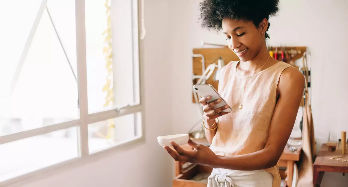 Uma mulher negra de blusa bege e calça branca está em pé segurand um objeto em uma mão enquanto tira uma foto dele com o celular. Ela está enviando a imagem de seu produto para um cliente por meio do WhatsApp Business