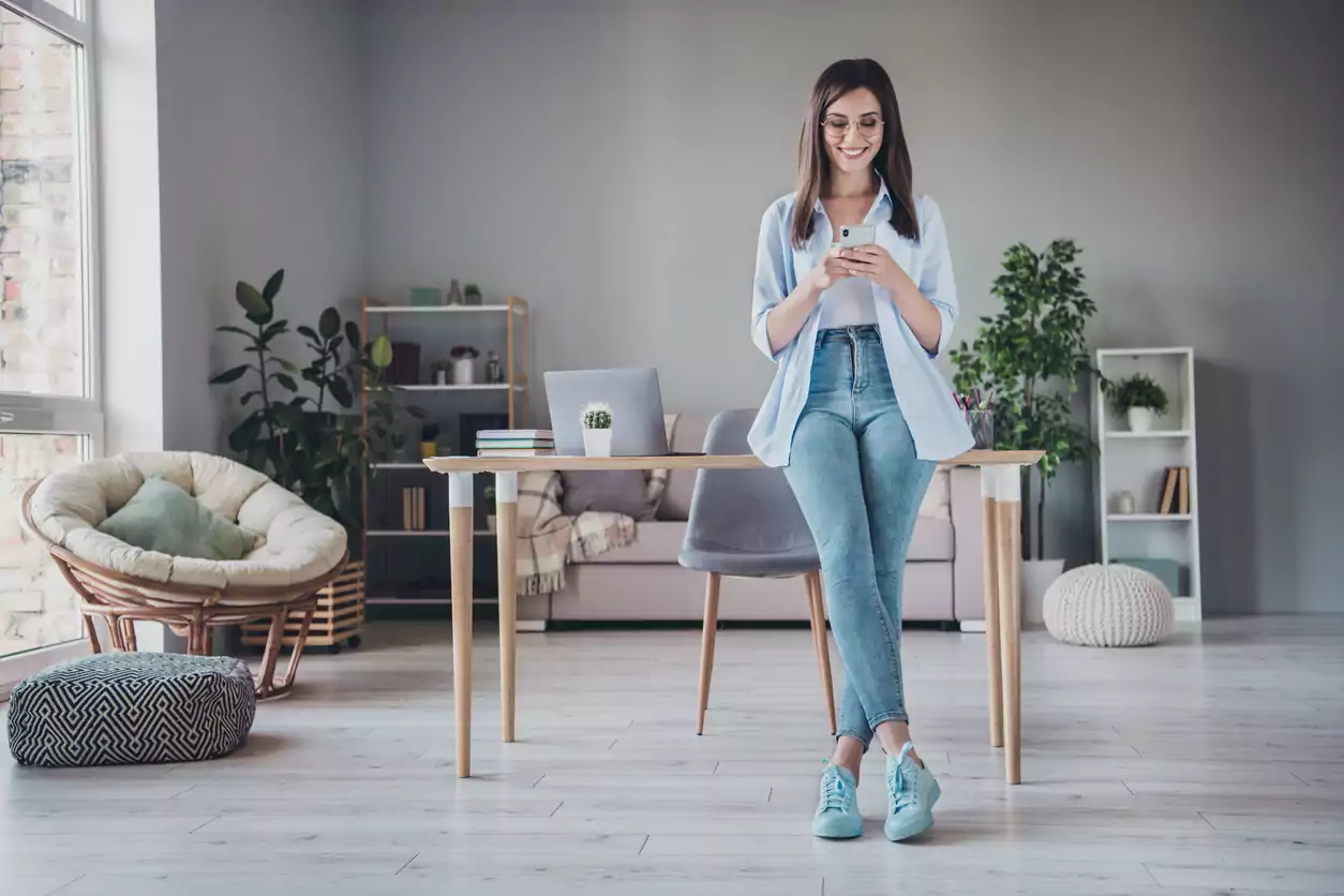 Uma mulher de cabelo liso, jaqueta azul clara, calça jeans azul e tênis azul está em pé em uma sala e apoiada em uma mesa. Ela segura um celular em suas mãos e olha para a tela sorrindo enquanto aprende os passos para construir uma boa presença digital para o seu negócio