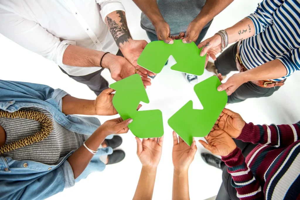 Grupo diversificado de mãos segurando o símbolo verde de reciclagem, representando a economia circular e a colaboração.