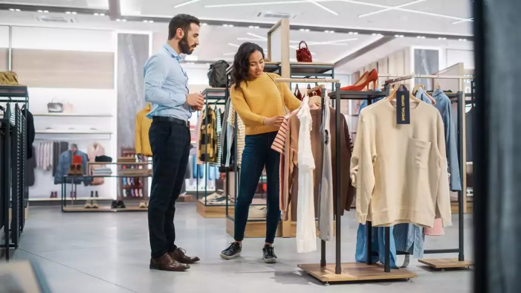 Uma mulher morena, de cabelo castanho, blusa de linha amarela de manga comprida, calça jeans escura e tenis preto está em uma loja olhando algumas roupas. Ela está sendo atendida por um homem branco, de cabelo e barba preta, blusa social azul clara, calça social xadrez e sapato social