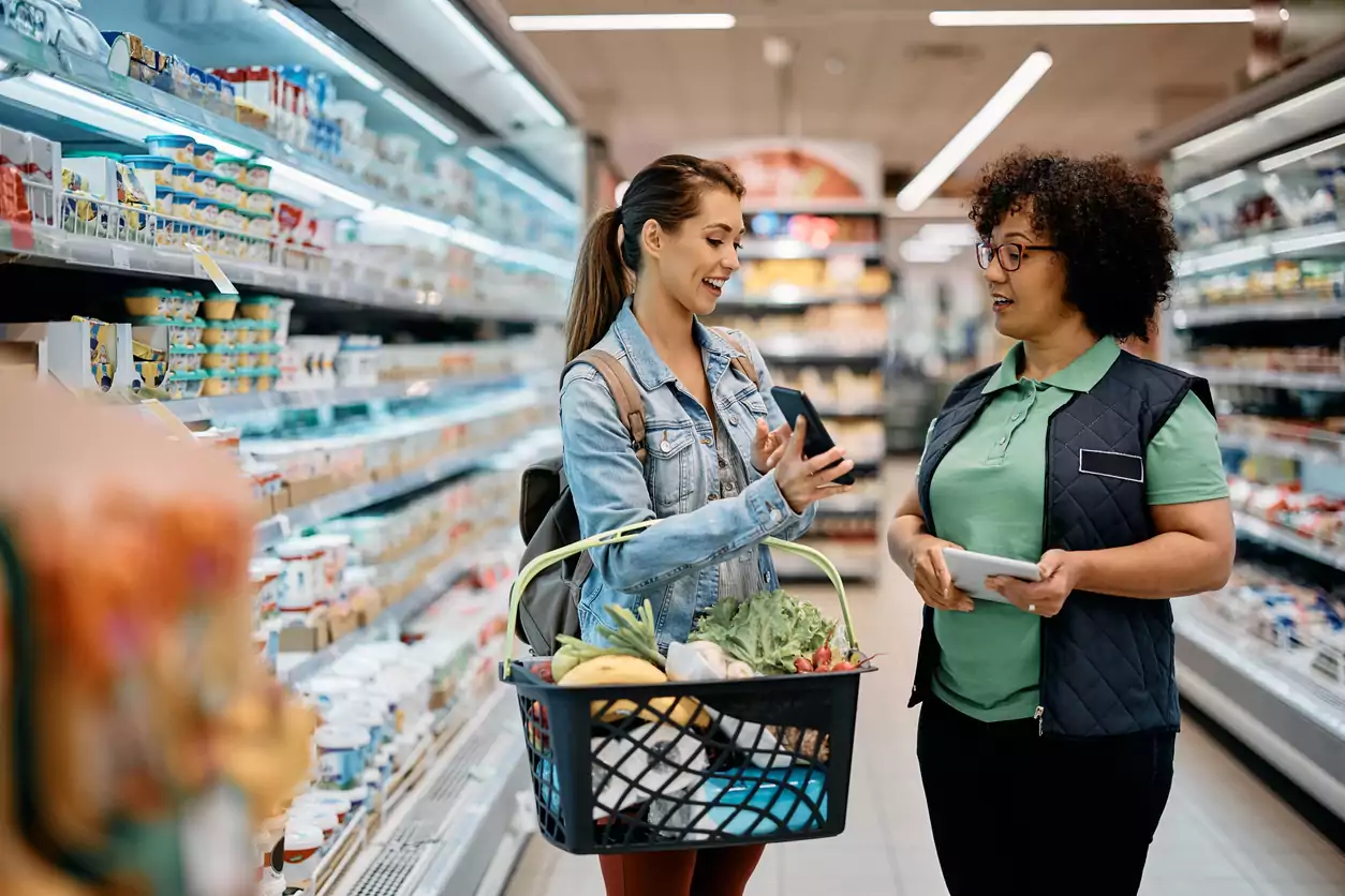 Uma mulher branca, de cabelo castanho claro liso e grande, preso em um rabo de cavalo, está usando uma jaqueta jeans, calça jeans marrom e uma mochila nas costas enquanto segura uma cesta de supermercado com varias compras. Ela está em pé mostrando o celular para uma vendedora, uma mulher morena, de cabelo cacheado preto curto, blusa polo verde claro, colete e calça preta. A antendente está segurando um bloquinho anotando os pedidos da cliente, a fim de colocar melhorar o customer experiencie oferecido pelo supermercado
