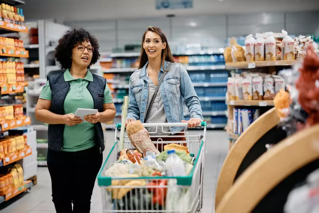 Uma mulher branca, de cabelo castanho claro liso grande, está usando uma blusa cinza e uma jaqueta jeans por cima. Ela está em um supermercado empurrando um carrinho com várias compras enquanto é acompanhada por uma atendente. Essa antendente é uma mulher morena, de cabelo preto cacheado, usando uma blusa polo verde claro, colete preto por cima e uma calça preta. a antendente está colocando em prática o que aprendeu sobre customer experience para ajudar a cliente
