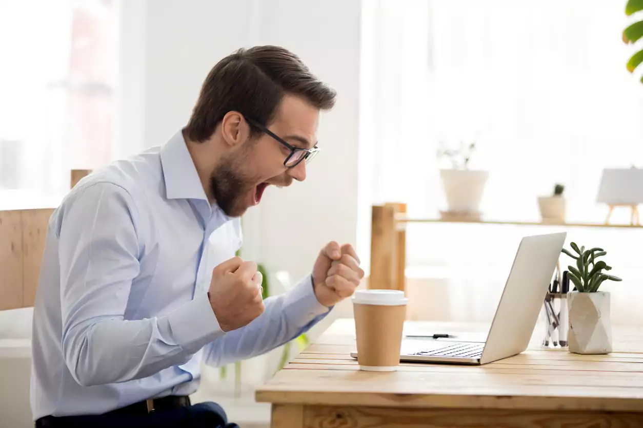 Um homem brano, de cabelo castanho curto e barba curta, está usando uma camisa social azul clara e um óculos de grau com armação preta. Ele está sentado em uma mesa de frente para um notebook e um copo de café. Ele está feliz, comemorando porque obteve sucesso ao aplicar tecnicas para analise de concorrencia