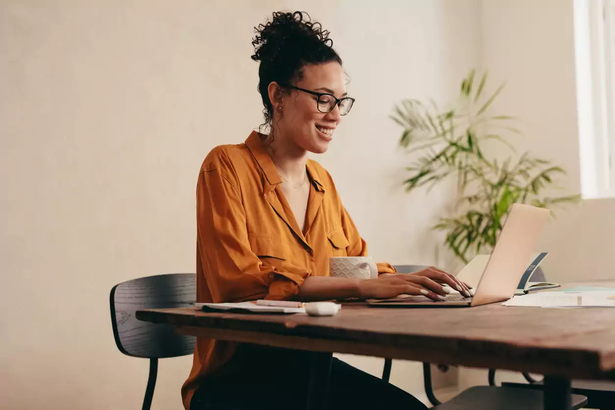 Uma mulher morena, de cabelo castanho escuro cacheado, que está preso e um coque, está usando uma blusa laranja de manga comprida e um óculos de grau. Ela está sentada em uma mesa, mexendo no notebook. Ela está sorrindo porque descobriu as principais tendências para o varejo em 2023 e vai utilizá-las em seu negócio