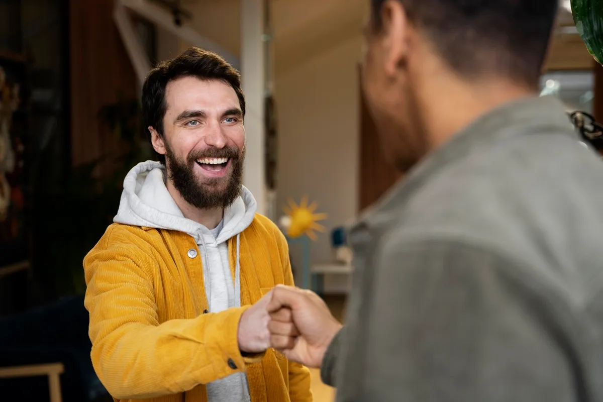 Homem sorridente cumprimentando outro com um aperto de mão, ambos vestindo roupas casuais em um ambiente interno iluminado.