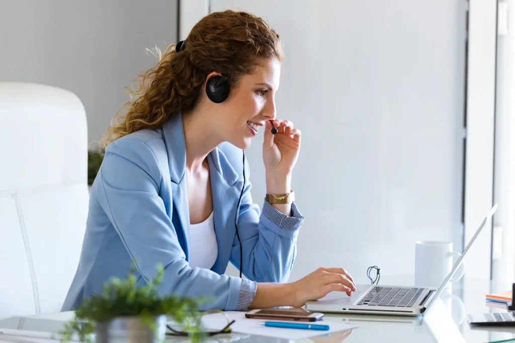 Mulher sorridente com fone de ouvido e microfone trabalhando em notebook, prestando atendimento ao cliente em ambiente claro e moderno.