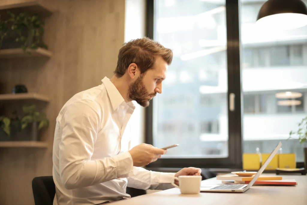 a imagem mostra um homem analisando informações em seu notebook, que está sobre a mesa com uma xícara de café ao lado, enquanto segura seu smartphone no escritório.