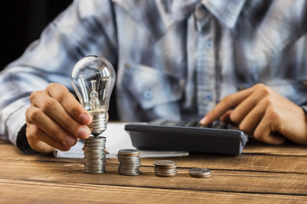 pessoa segurando uma lâmpada em cima de várias moedas sobre a mesa enquanto mexe em uma calculadora
