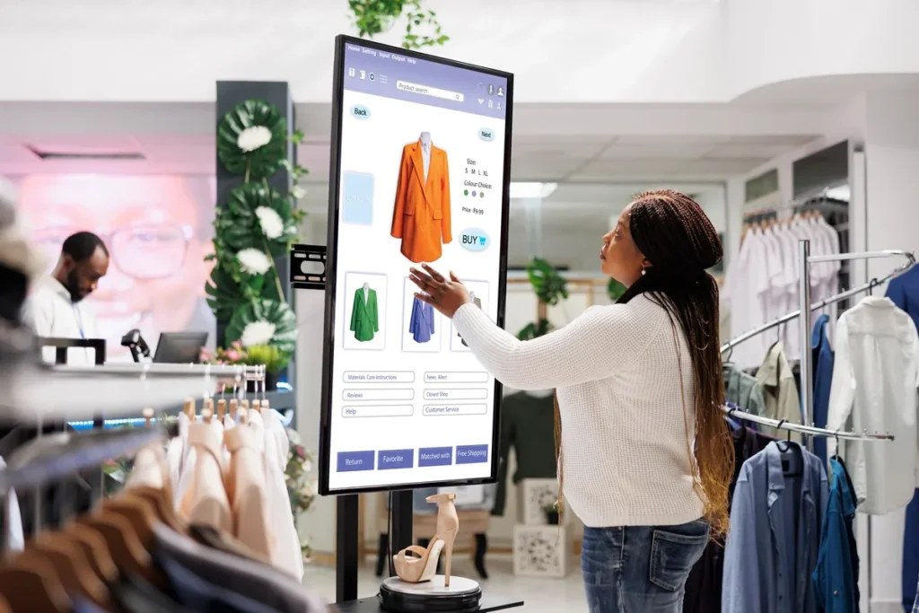 Mulher interagindo com tela digital em loja de roupas, navegando em opções de casacos disponíveis em cores e tamanhos, simbolizando inovação e experiência de compra omnichannel.