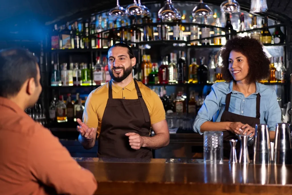Bartenders sorridentes atendendo um cliente em um bar sofisticado, destacando um ambiente acolhedor e de excelência no atendimento.