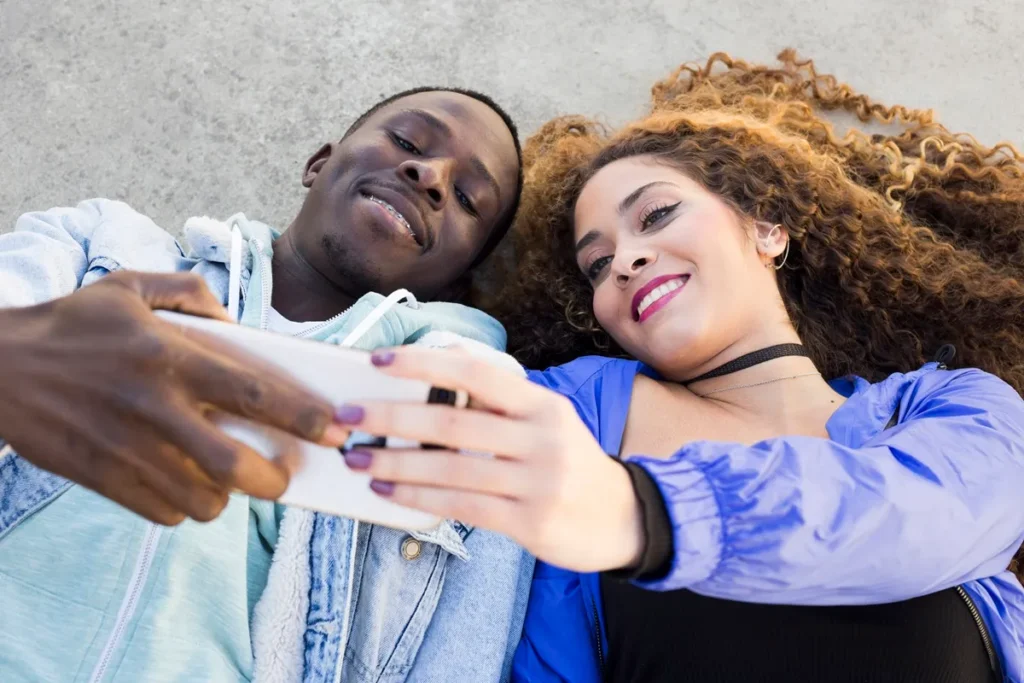 Casal jovem tirando uma selfie com smartphone, deitado no chão ao ar livre, sorrindo e aproveitando um momento descontraído, representando a conexão e autenticidade da geração Z.