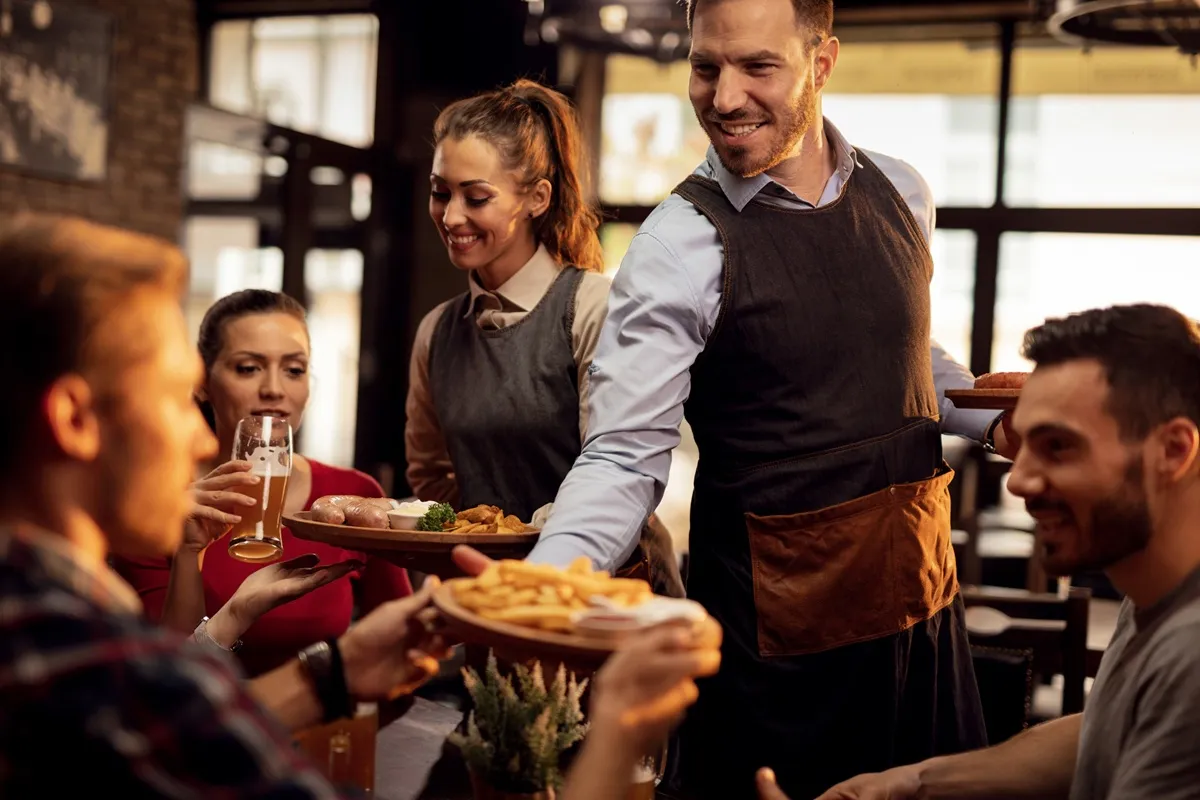 Garçons sorridentes servindo comida e bebida a clientes felizes em um restaurante sofisticado, destacando a qualidade do atendimento.