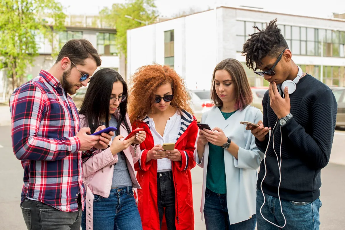 Grupo de cinco jovens adultos, em ambiente urbano, concentrados em seus smartphones, representando o uso intenso de tecnologia e o comportamento digital característico da geração Z