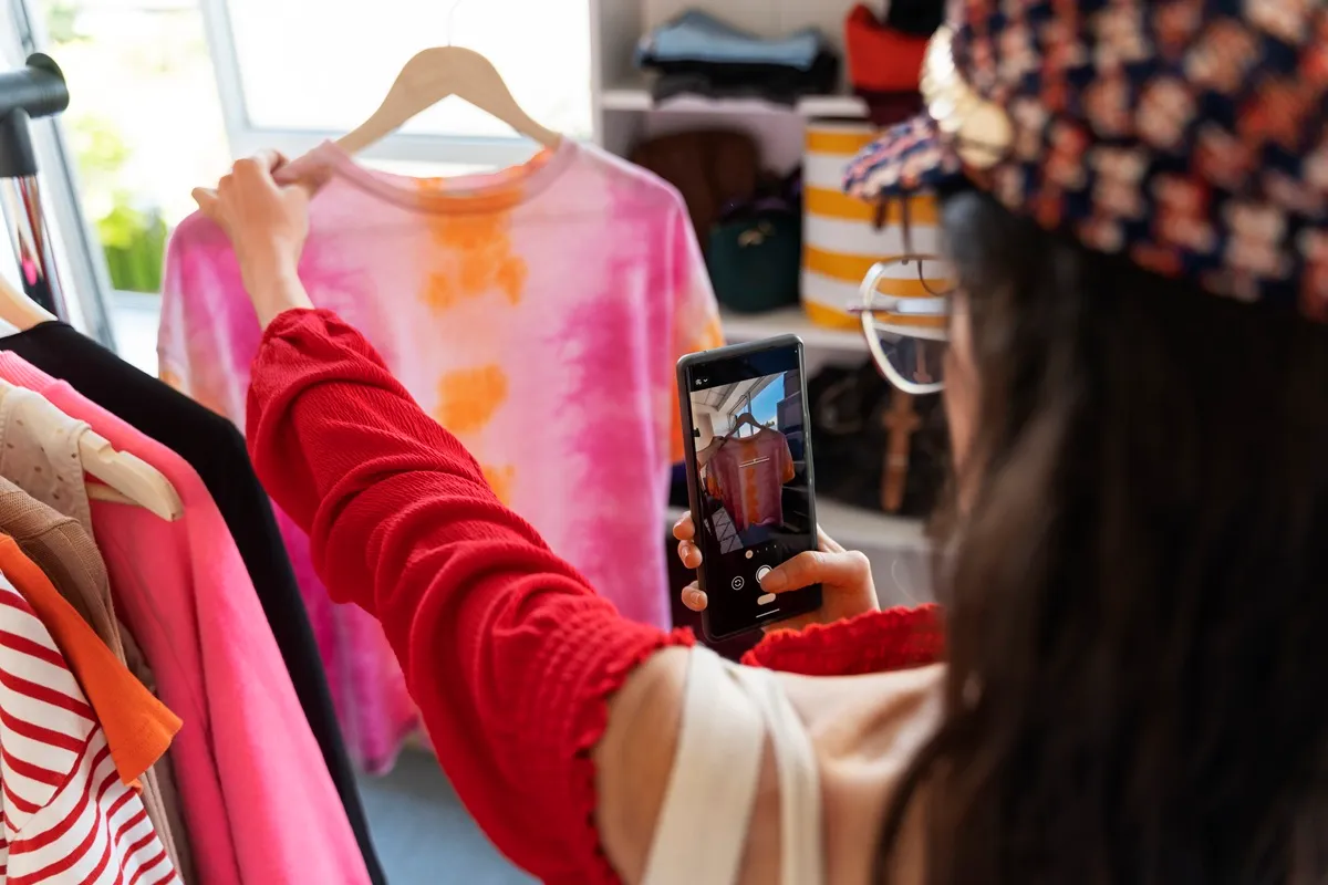 Mulher segurando camiseta tie-dye rosa e laranja enquanto fotografa com seu smartphone em uma loja de roupas, representando compartilhamento de tendências de moda em redes sociais