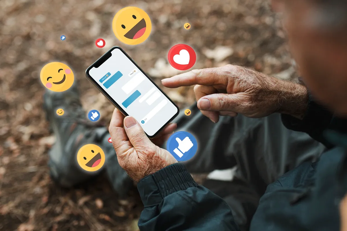 Idoso utilizando smartphone para interagir em redes sociais, com emojis flutuando ao redor, representando curtidas e reações.