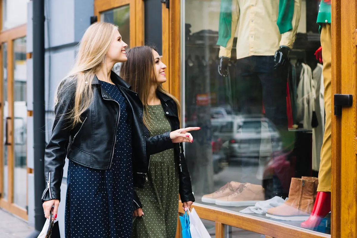 Duas mulheres jovens com sacolas de compras observando a vitrine de uma loja com roupas e calçados em exposição, sorrindo e apontando para os itens.