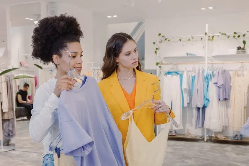 Duas mulheres numa loja de roupa, uma delas a segurar uma camisola para a outra, que está a usar um blazer amarelo. A imagem ilustra a experiência de compra personalizada e a importância do serviço para o CLV.