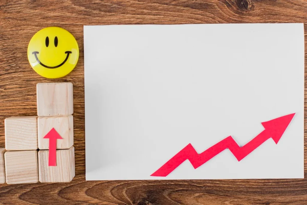 Seta vermelha de crescimento sobre papel branco, ao lado de blocos de madeira com seta para cima e botão amarelo com carinha feliz, sobre mesa de madeira.