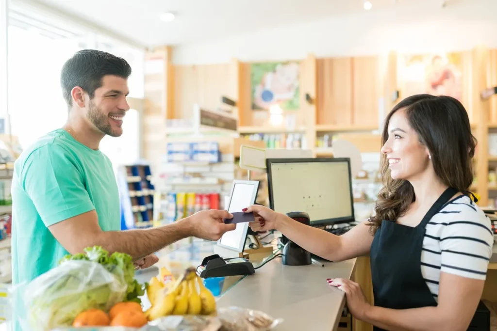 Funcionária de supermercado com avental preto sorrindo enquanto recebe pagamento com cartão de crédito de um cliente no caixa.