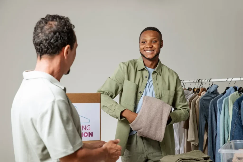 Homem sorridente segurando uma peça de roupa enquanto participa de uma campanha de doação, com um voluntário à sua frente e um cabideiro cheio de roupas ao fundo.