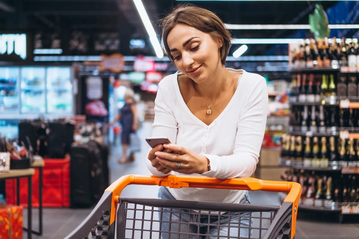 Mulher sorridente encostada em um carrinho de compras no supermercado, usando o celular enquanto faz compras.
