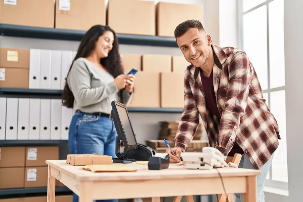 Empresário sorridente organiza inventário em um pequeno armazém, enquanto uma funcionária confere informações no celular, representando controle de estoque eficiente.