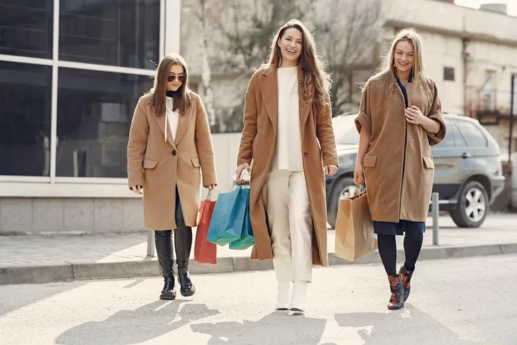 Três mulheres sorridentes, com casacos de inverno, carregando sacolas de compras em uma calçada, simbolizando as ofertas da temporada.