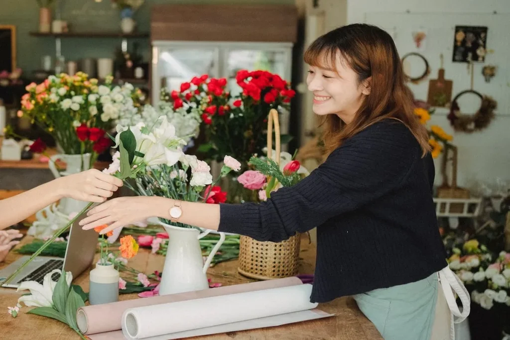 Florista com um sorriso, passando uma flor branca para uma cliente, em uma loja com buquês coloridos, exemplificando organização.