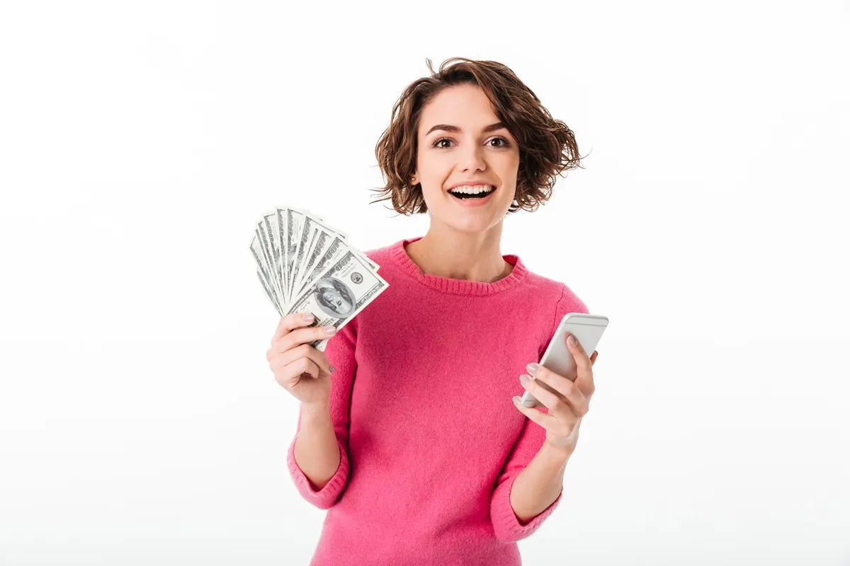 Mulher feliz segurando um maço de notas de dólares em uma mão e um celular na outra, sorrindo para a câmera.