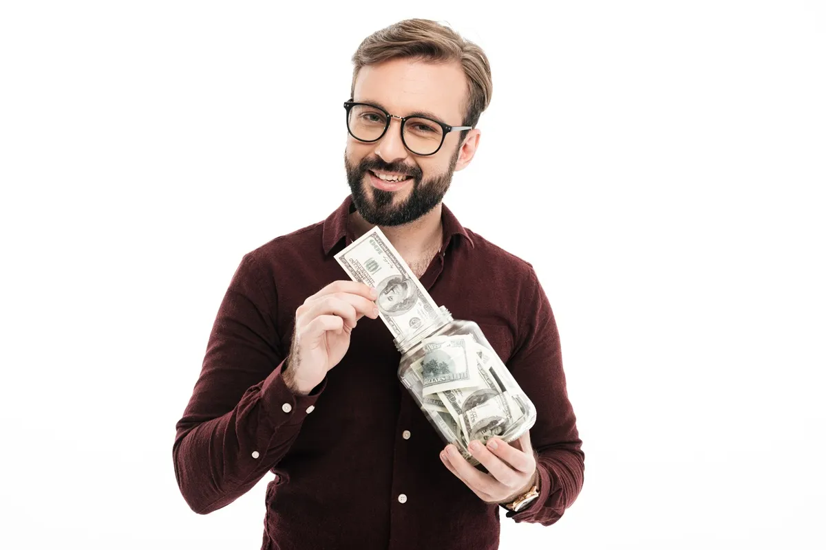 Homem sorrindo enquanto coloca notas de 100 dólares em um pote, com uma expressão de satisfação.