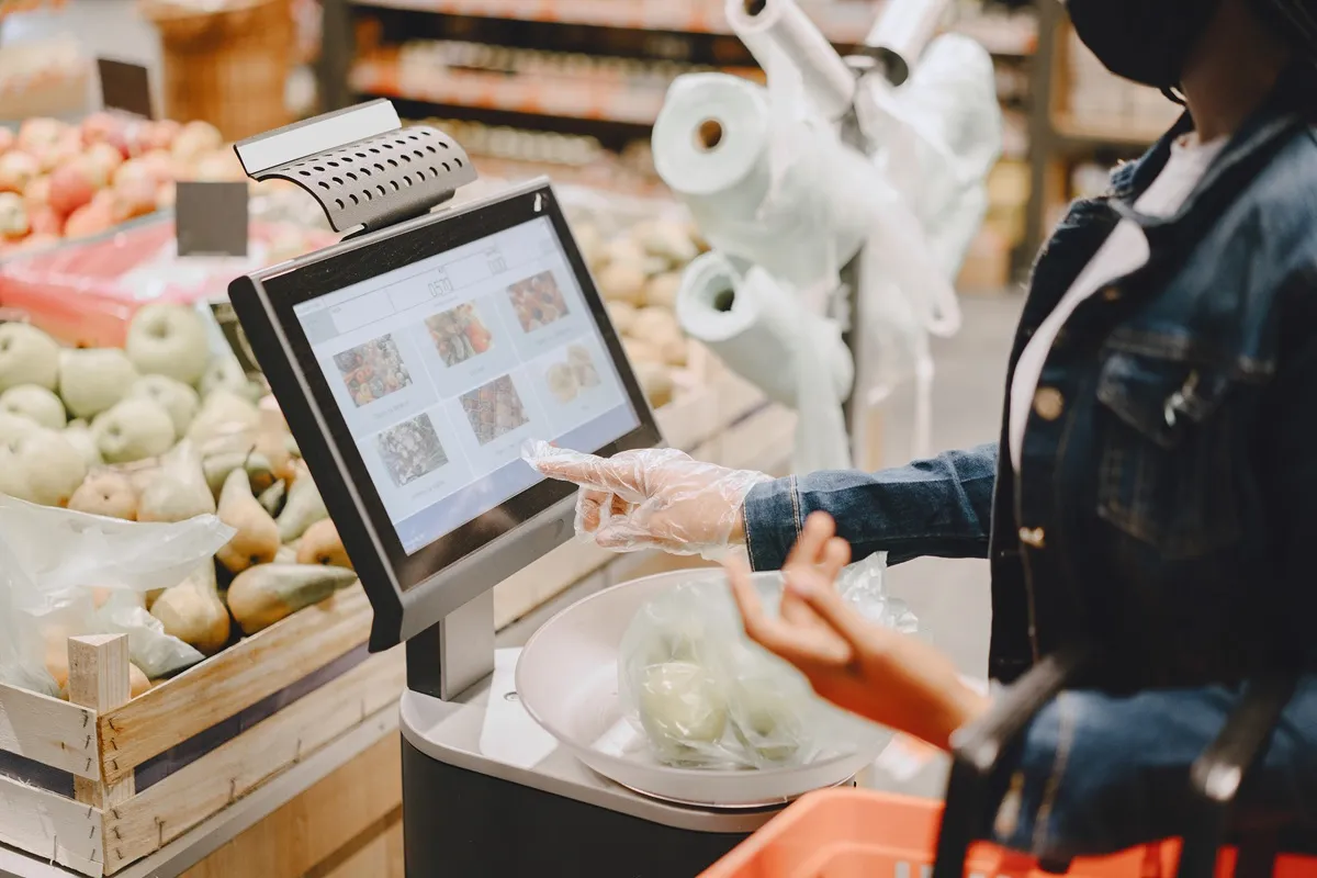 Pessoa usando luva plástica toca em tela de balança digital para pesar frutas no setor de hortifrúti do supermercado.