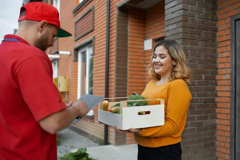 Entregador com uniforme vermelho, segurando um tablet, entregando uma caixa de vegetais e frutas para uma cliente, simbolizando o serviço de delivery.