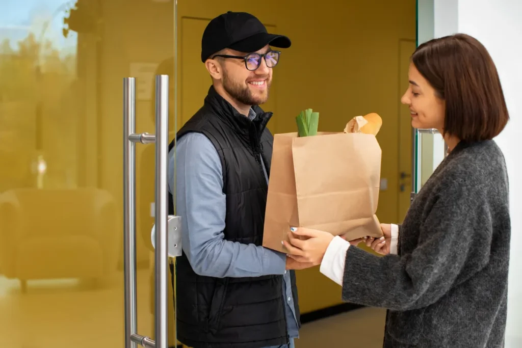 Entregador de boné e óculos sorrindo, entregando uma sacola de papel com compras para uma cliente, ilustrando o serviço de delivery.