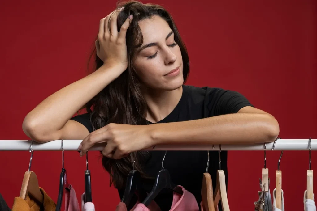Mulher com expressão pensativa encostada em arara de roupas, indecisa sobre o que vestir, com fundo vermelho