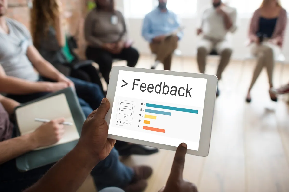 Pessoa segurando um tablet com uma tela exibindo feedback com barras de avaliação em estrelas e gráficos coloridos, enquanto um grupo de pessoas está ao fundo.